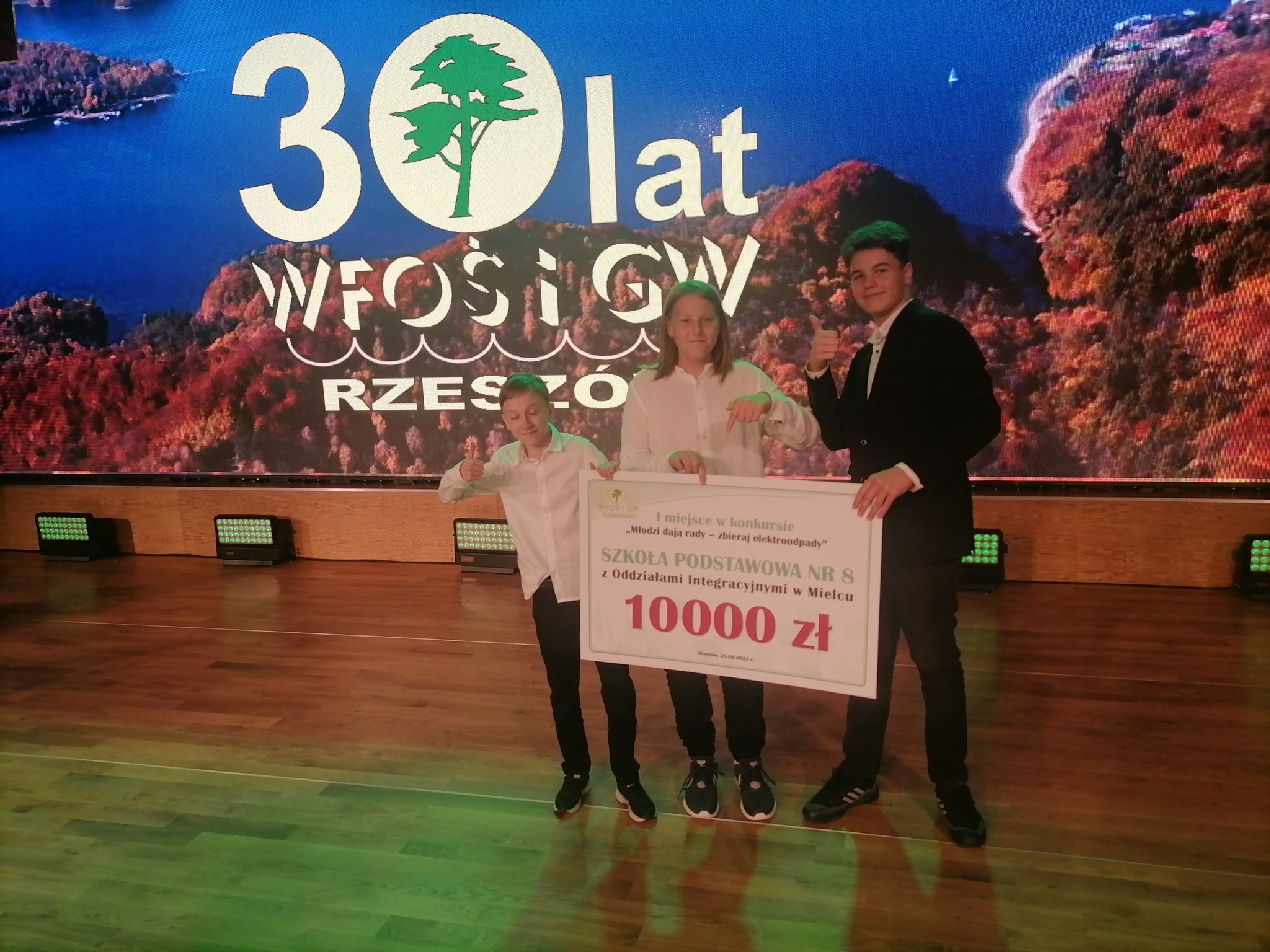 Zdjęcie przedstawia trójkę uczniów SP8. Pozują oni z czekiem na sumę 10000 złotych - nagrodą w konkursie. Uczniowie uśmiechają się i wyrażają zadowolenie z wygranej w konkursie.