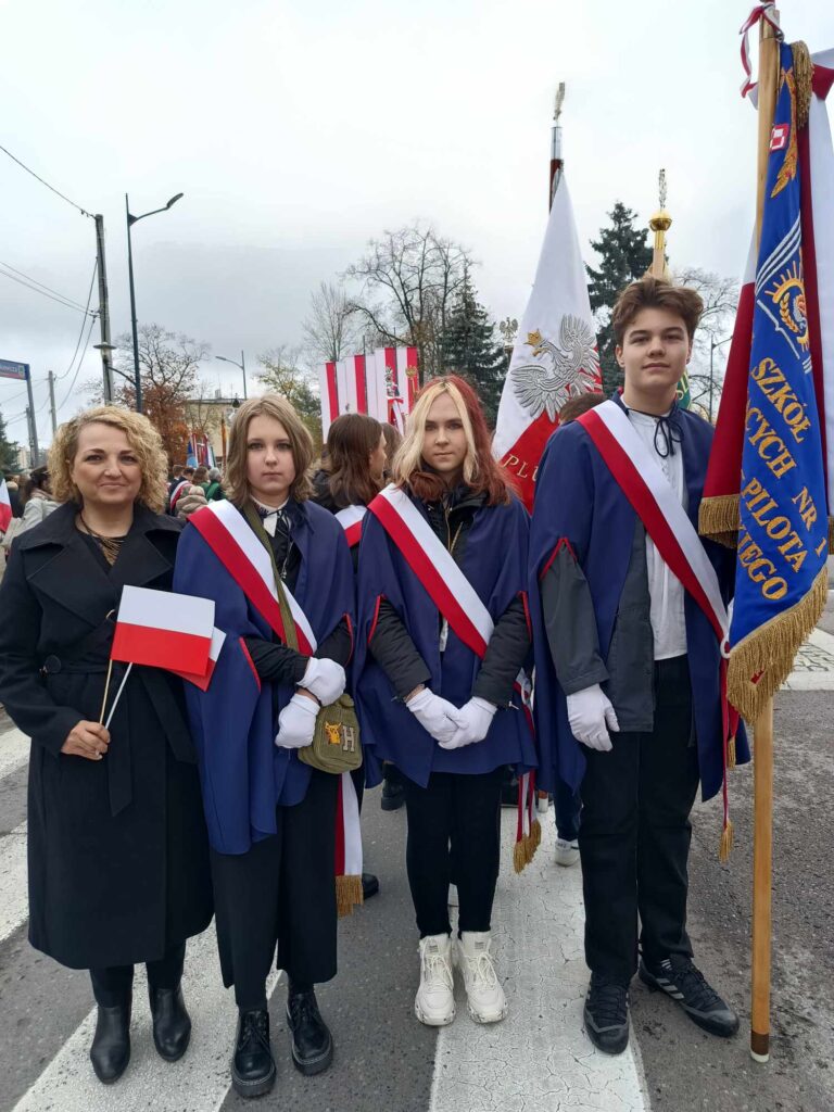 Zdjęcie przedstawia p. dyrektor szkoły, Joannę Gładyszewską, razem z uczniami z pocztu sztandarowego. Uczniowie mają biało-czerwone szarfy, p. dyrektor trzyma w ręku flagę narodową.