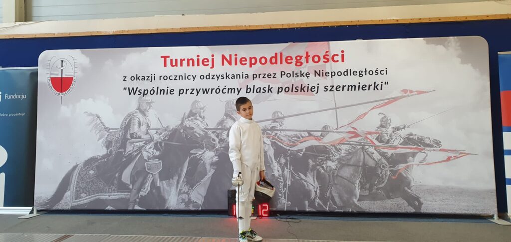 Na zdjęciu Olek Murdza w stroju szermierczym. Olek pozuje na tle plakatu promującego Turniej NIepodległości