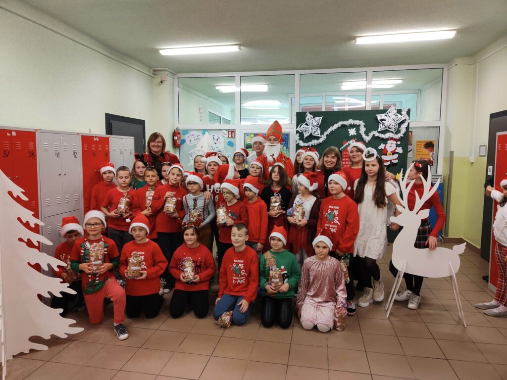 Uczniowie na zdjęciu są ubrani w czerwone świąteczne stroje, wszyscy mają czapki św. Mikołaja. W rękach trzymają prezenty otrzymane od św. Mikołaja