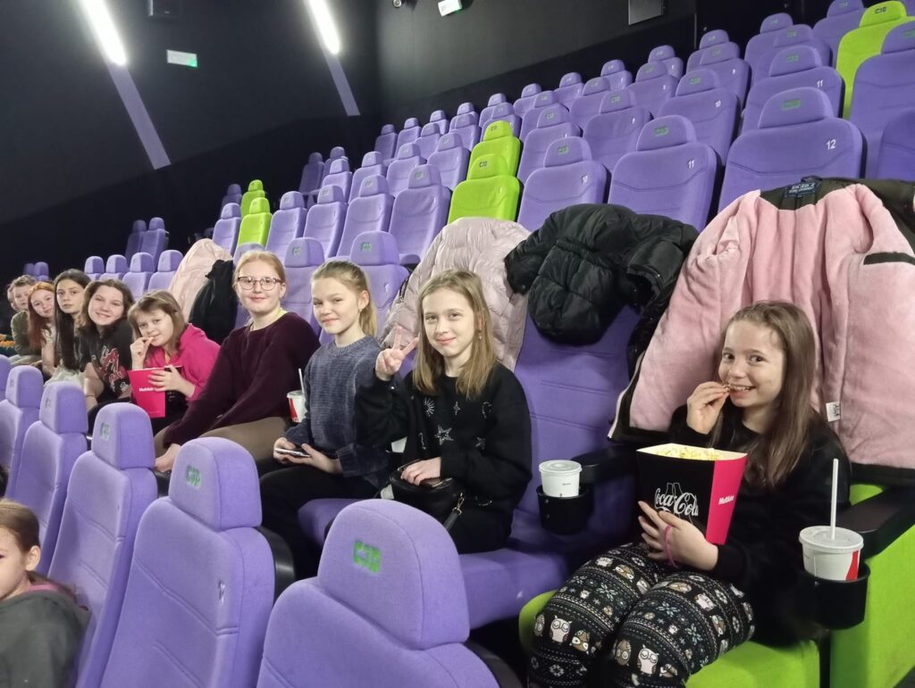 Zdjęcie przedstawia uczennice klasy czwartej na sali kinowej. Uczennice siedzą w jednym rzędzie, niektóre  trzymają w rękach duże opakowania popcornu. Wszyscy uśmiechają się do zdjęcia.