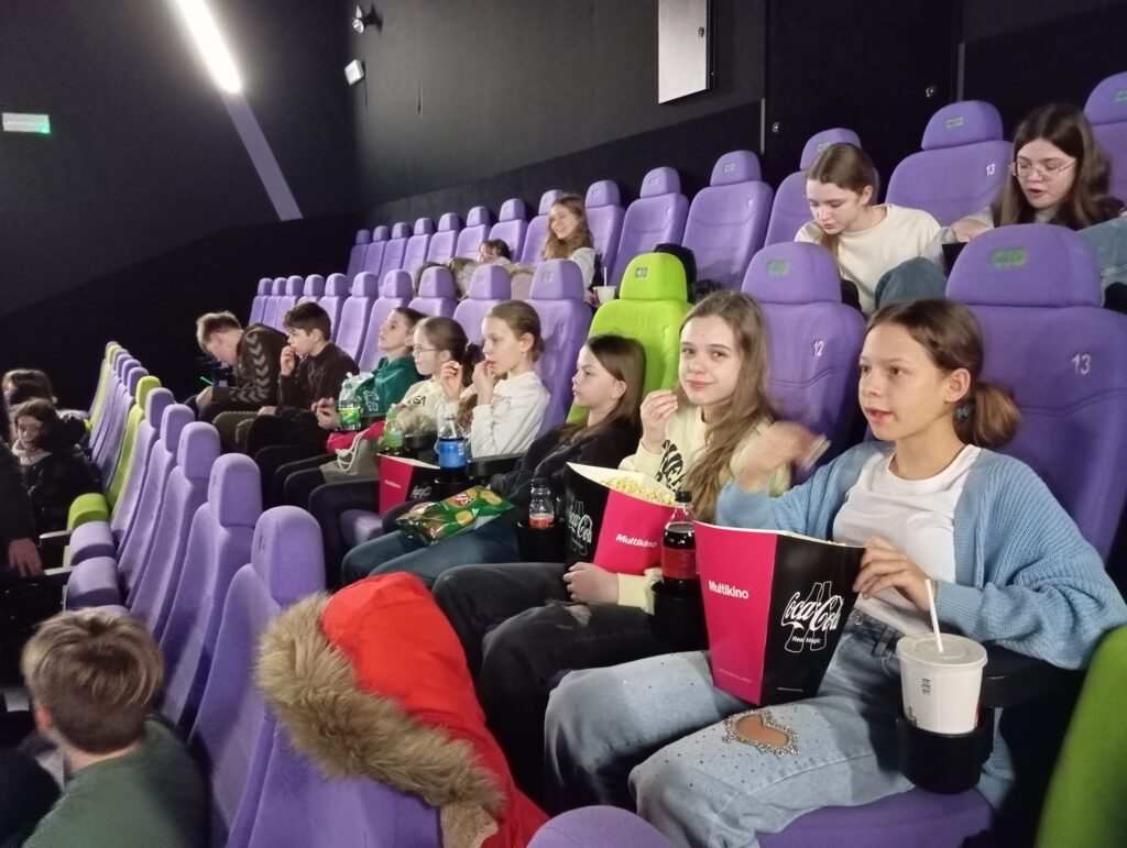 Zdjęcie przedstawia grupę uczniów Sp8 oczekujących w kinie na rozpoczęcie filmu. Kilka osób na zdjęcia trzyma w rękach nieodłączny atrybut kinomaniaka - duże pudło popcornu.
