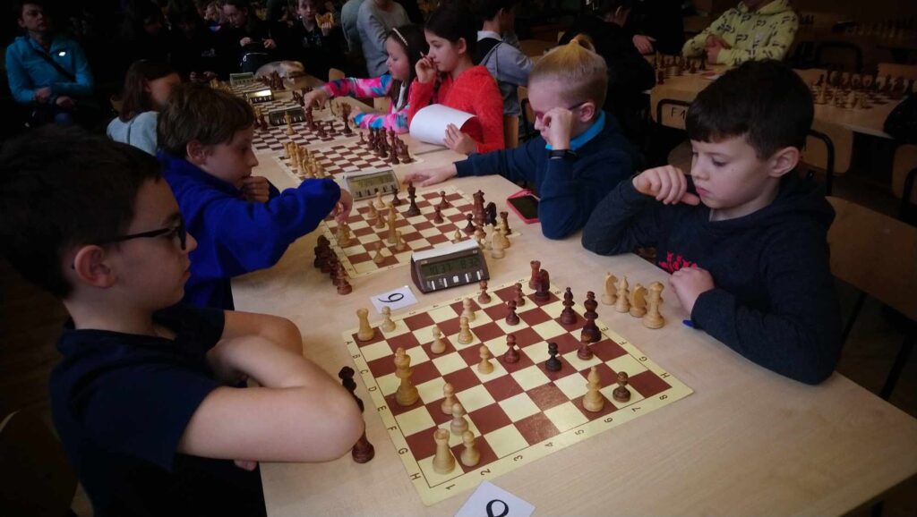 Zdjęcie przedstawia uczniów grających w szachy. Na stoliku rozstawiona jest szachownica, uczniowie obserwują ją w skupieniu. Na drugim planie podobna sytuacja: także rozgrywany jest mecz szachowy.