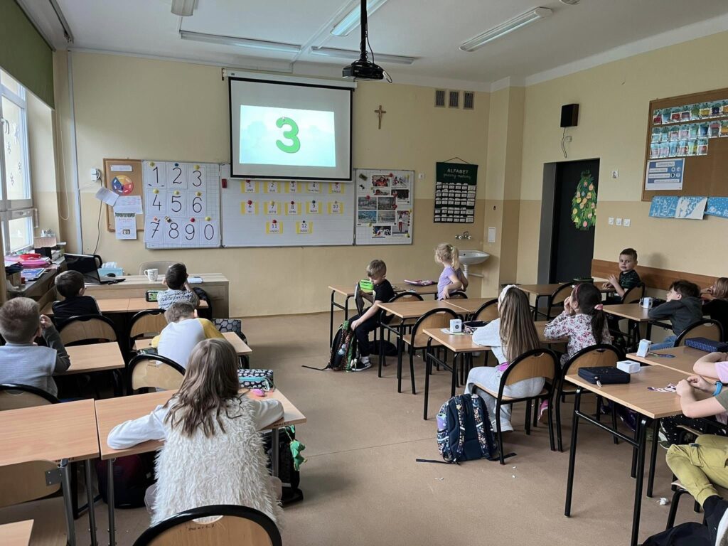 Zdjęcie przedstawia uczniów klasy 1c, uczących się liczyć po włosku. Uczniowie siedzą przed tablicą, na której przedstawione są liczby 0-9 zapisane po włosku, nauka odbywa się także przy zastosowaniu rzutnika.